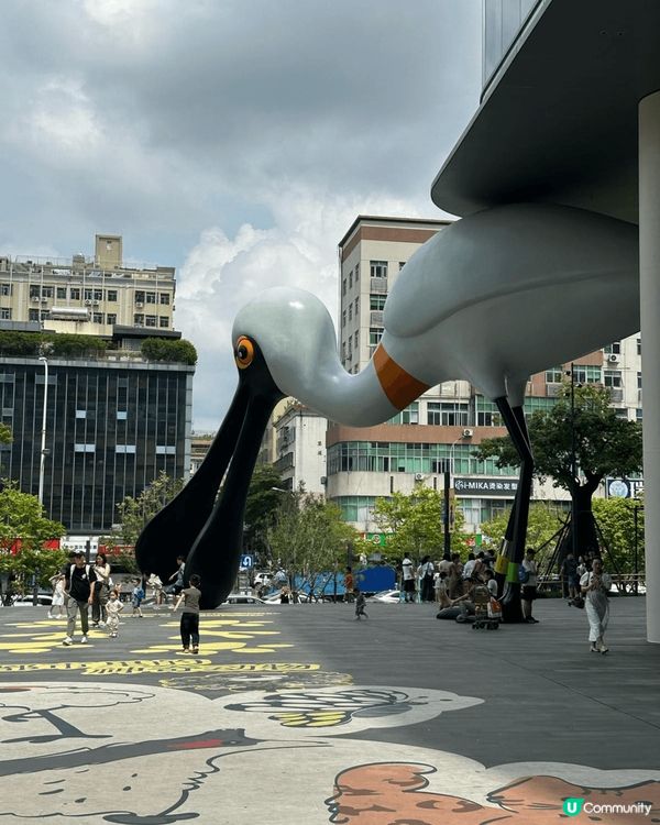 這張圖片是一個戶外廣場，有一隻超巨型的白色鳥雕塑，鳥嘴很長，...