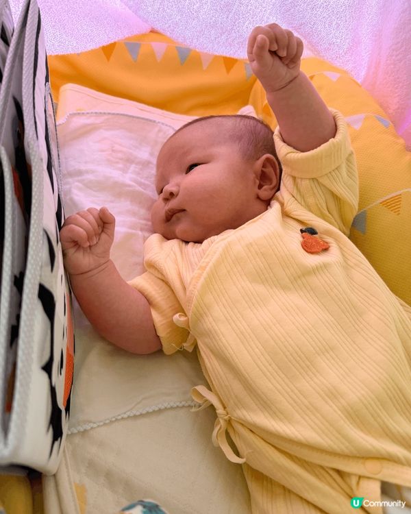 BB躺在柔軟的黃色嬰兒床中，雙手自然舉起，像是在伸展或開心地...